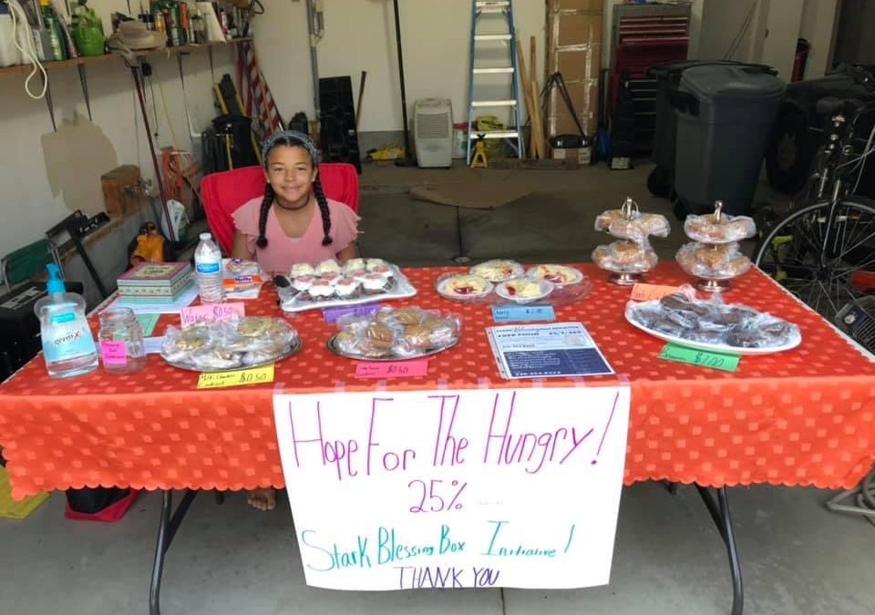 Laura sits at a table. Packaged baked goods sit on the table. A large posterboard sign is affixed to the table and reads 'Hope for the Hungry 25% Stark Blessing Box Initiative Thank You!'