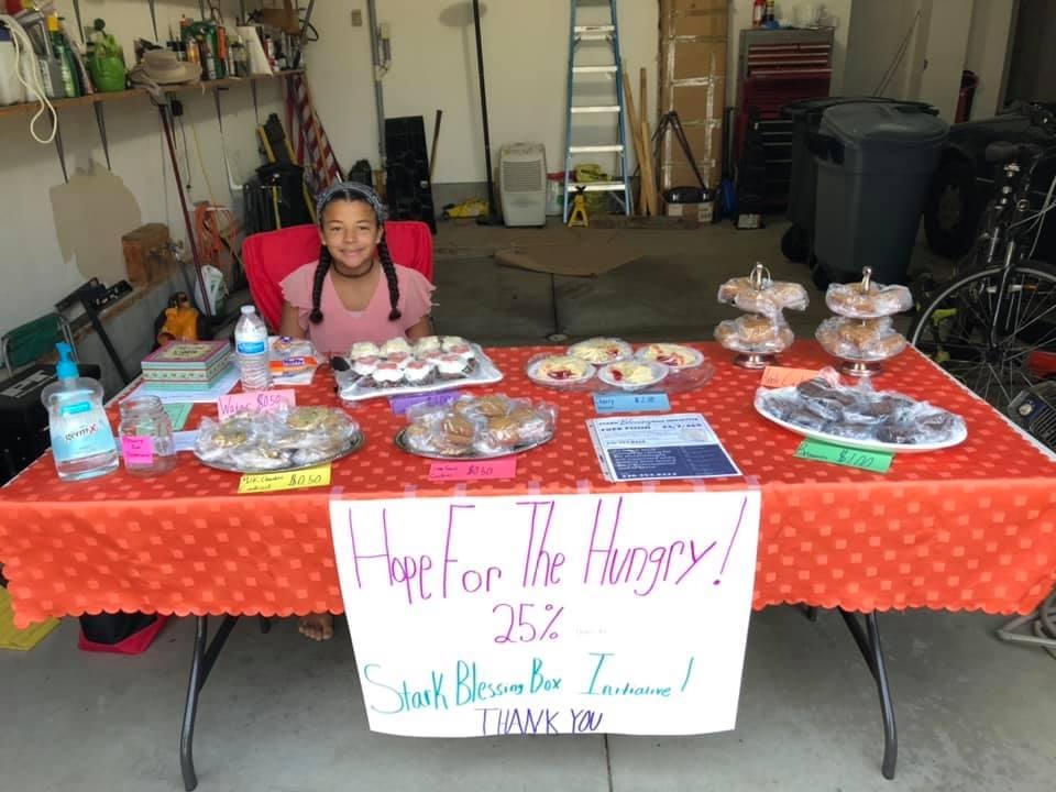 Laura sits at a table. Packaged baked goods sit on the table. A large posterboard sign is affixed to the table and reads 'Hope for the Hungry 25% Stark Blessing Box Initiative Thank You!'